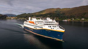 Havila Kystruten skip som kjører på fjorden, med fjell og overskyet himmel i bakgrunnen.